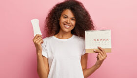 Portrait of good looking feminine girl has Afro hairstyle, holds monthly calendar, clean sanitary napkin, has menstrual cycle, happy to have good women health. People, gynecology, hygiene concept